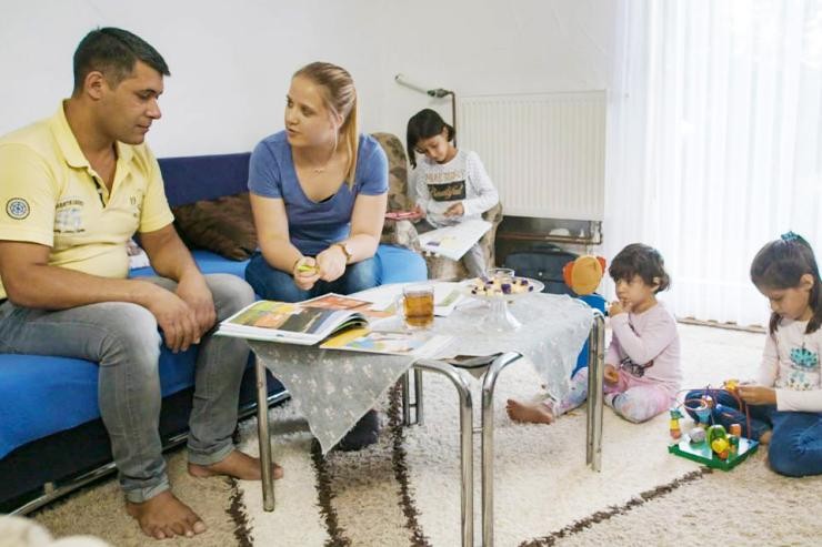 Studierende berät einen Familienvater in dessen Wohnung, Kinder spielen im Hintergrund